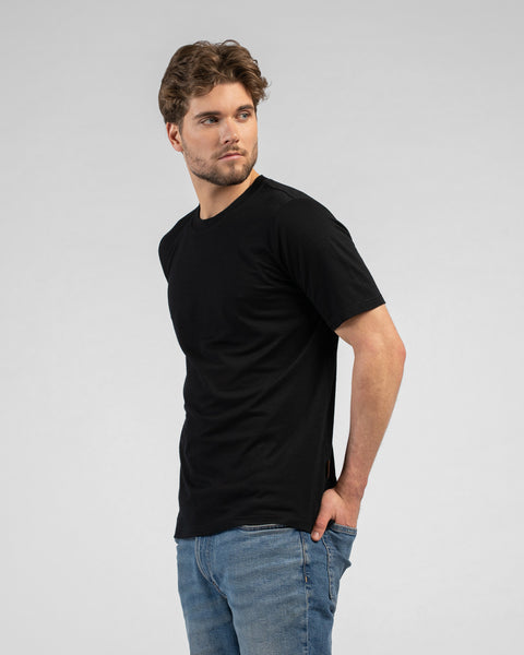 A man stands facing away but looks back over his left shoulder. He wears a black short-sleeved t-shirt and blue jeans, with his left hand casually tucked into his back pocket.