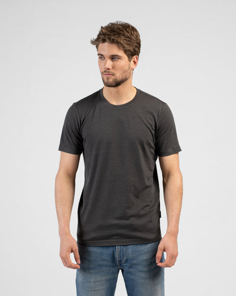 A studio portrait of a man standing and looking off to the left side of the frame against a plain white background. He is wearing a fitted, charcoal grey short-sleeved crew-neck t-shirt and blue jeans.