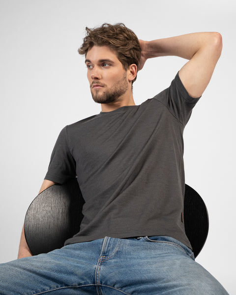 A man sits casually backward on a black chair, looking off to the left with one hand resting behind his head. He is wearing a charcoal grey short-sleeved t-shirt and blue jeans.