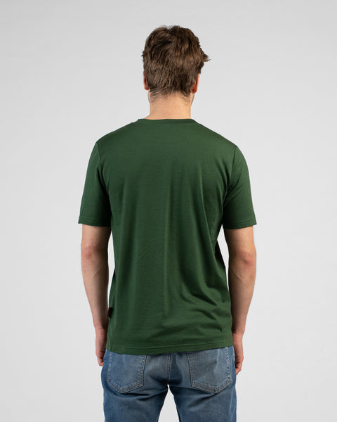 A studio shot showing the back of a man wearing a dark green short-sleeved t-shirt and blue jeans. He stands facing away from the camera against a plain light grey background.