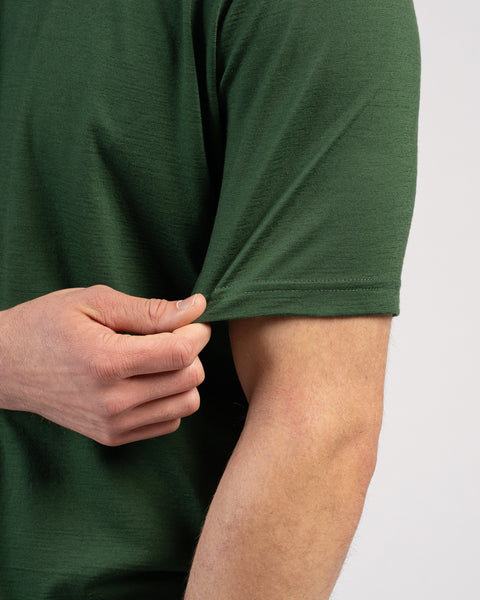A close-up shot of a man wearing a dark green short-sleeved t-shirt against a plain light grey background. His hand is gently pinching and pulling the hem of the sleeve, highlighting the fabric's texture and flexibility.