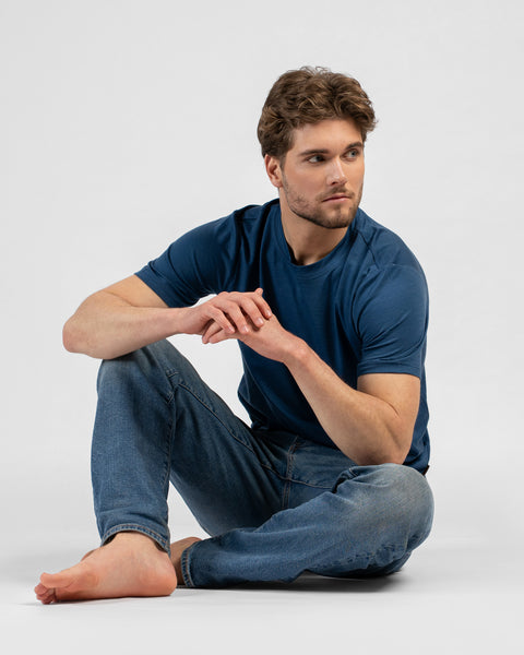 A barefoot man sits on the floor, leaning casually with one arm resting on a black stool while looking to the right. He is wearing a denim blue short-sleeved t-shirt and blue jeans.