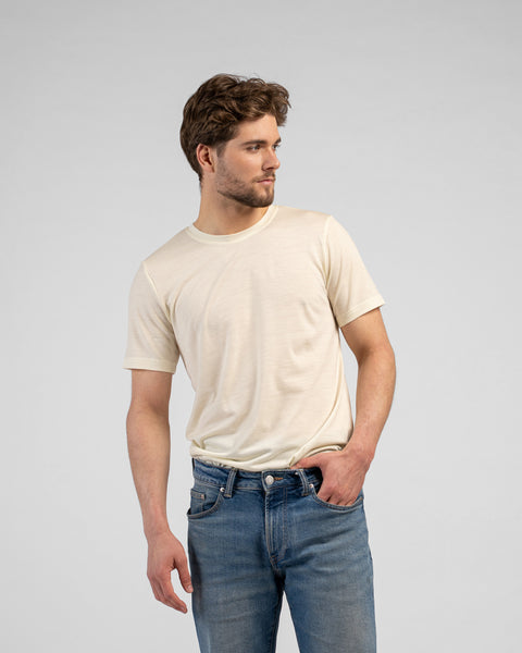 A studio portrait of a man standing and looking off to the right side of the frame against a plain white background. He has his left hand casually resting in the pocket of his blue jeans and is wearing a fitted, light natural cream-colored short-sleeved crew-neck t-shirt.