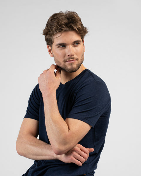 A side-profile studio portrait of a man looking off to the right. He is wearing a dark navy blue short-sleeved crew-neck t-shirt with his hand thoughtfully resting on his chin.