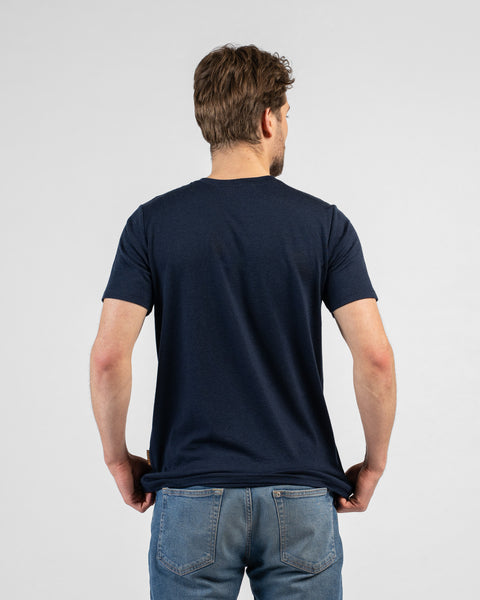 A studio shot showing the back of a man wearing a dark navy blue short-sleeved crew-neck t-shirt and blue jeans. He stands facing away from the camera, holding the bottom hem of his shirt.