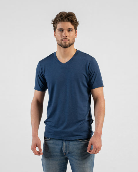 A studio portrait of a man looking directly at the camera with his hands relaxed by his sides. He is wearing a denim blue v-neck t-shirt and blue jeans against a light grey background.