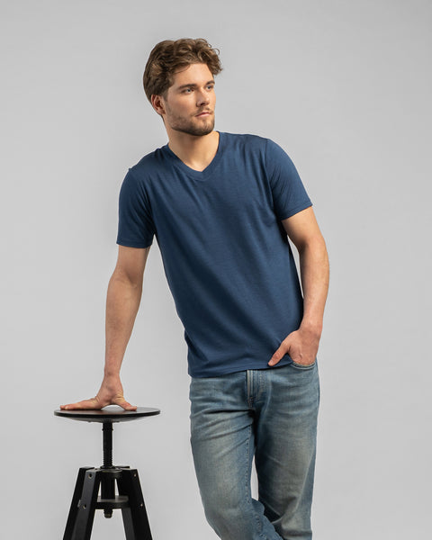 A man stands leaning casually with his right hand on a black stool, looking off to the right. He wears a denim blue v-neck t-shirt and blue jeans against a light grey background.