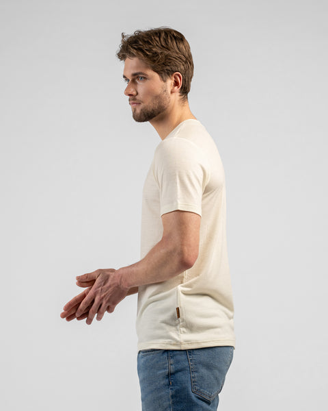 A side-profile shot of a man rubbing his hands together, looking to the left. He wears a light beige v-neck t-shirt and blue jeans against a light grey background.