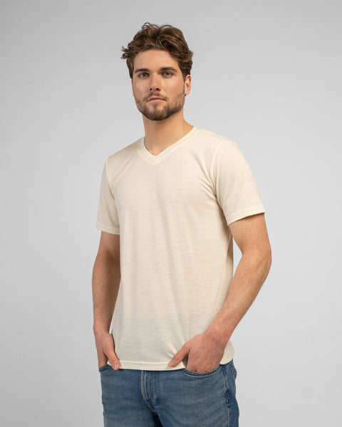 A studio portrait of a man standing and looking directly at the camera with his hands resting in his front pockets. He is wearing a light beige v-neck t-shirt and medium-wash blue jeans.