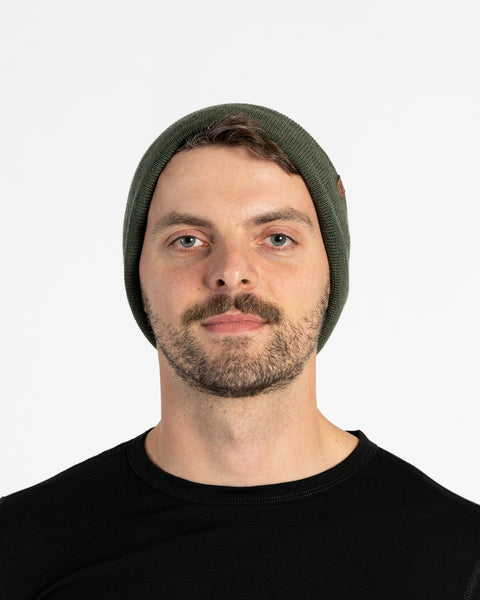 A person with light skin, short brown hair, a mustache, and light-colored eyes is shown in a headshot against a white background. They are wearing a dark green knit beanie with a small brown tag on the side and a black long-sleeved shirt. They are looking directly at the camera.