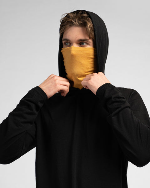 A studio portrait of a man looking off to the left. He is wearing a black long-sleeved hooded shirt with the hood up and a mustard yellow neck gaiter pulled up to cover his nose and mouth, adjusting the fabric with both hands.