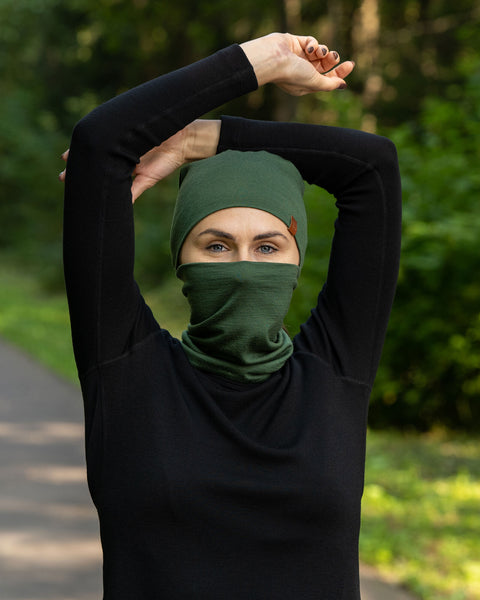 The picture features a woman wearing the unisex gaiter and beanie merino set in dark green color. She has her hands raised above her head. She is in a forest.