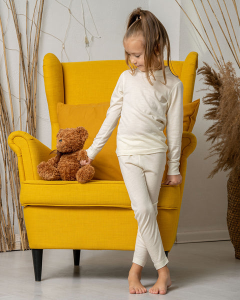A girl standing in front of a yellow armchair, wearing the menique long sleeve merino top in natural color and matching pants. She is holding a teddy bear by the hand, that is put on the armchair.
