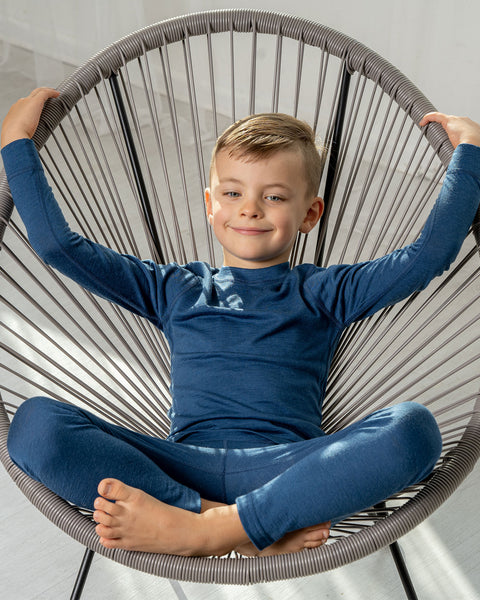 A boy wearing the menique kids' 160gsm merino long sleeve set in denim color. he is sitting in a lounge chair, with his legs crossed and hands raised.
