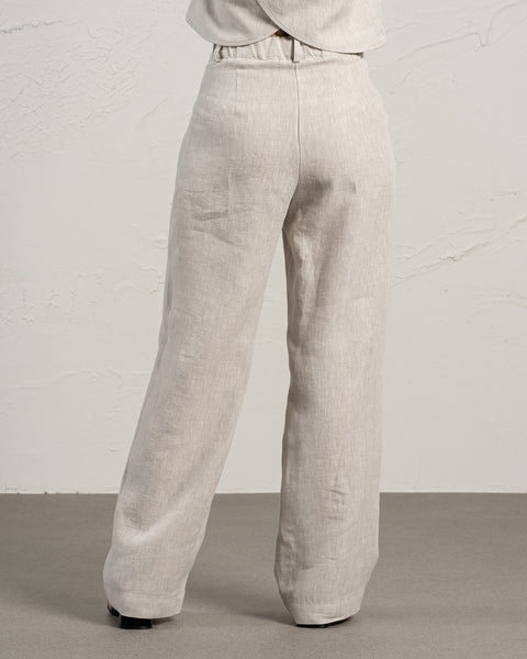 The photo shows the back view of a person wearing natural color, high-waisted, wide-legged linen menique pants. The trousers have belt loops and a slightly visible waistband. The person is wearing black shoes. The background consists of a textured light beige wall and a light gray floor.