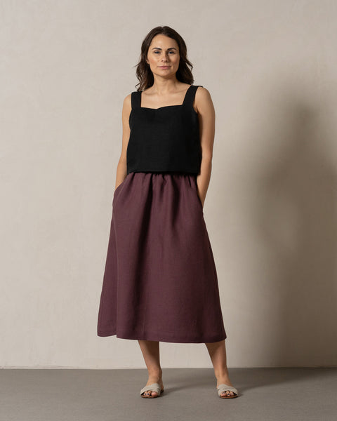 A woman with medium-toned skin and dark hair stands facing the camera against a neutral background. She is wearing a pure black, sleeveless square-neck linen top and a flowy, maxi skirt in a shadow purple color, with her hands casually placed in the skirt's pockets and light-colored sandals on her feet.