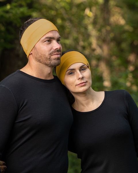 A man and a woman are standing close together outdoors, both wearing menique spicy yellow headbands and black long-sleeved shirts. The man has short brown hair and a beard, and he is looking slightly upwards and to the left.