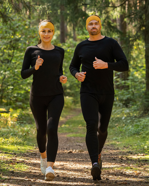 A man and a woman are jogging towards the viewer on a dirt path through a sun-dappled forest. They are both wearing spicy yellow headbands, black long-sleeved shirts, and black leggings, suggesting they might be a couple exercising together.
