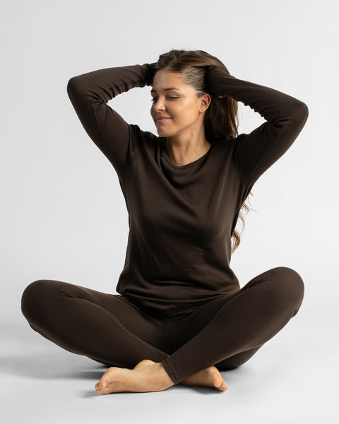 A barefoot woman sitting cross-legged on the floor with her hands behind her head, wearing a dark brown long-sleeve thermal lounge set against a light background.