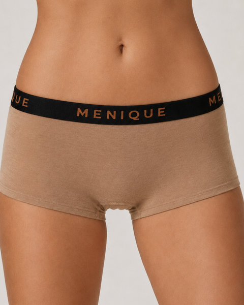Close-up studio photo of a woman wearing low-waist beige short boxer briefs with a black MENIQUE logo waistband, cropped from waist to upper thighs.