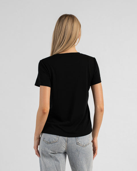 A back view studio shot of a blonde woman standing against a plain light grey background. She is wearing a black short-sleeved crew neck t-shirt and light wash denim jeans, with her straight blonde hair falling down her back.
