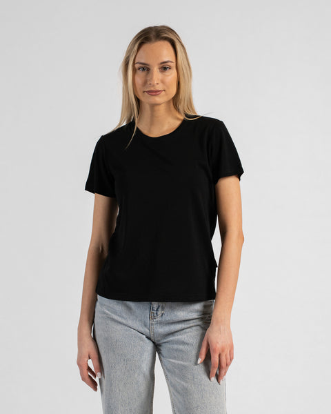 A studio portrait of a blonde woman standing and looking directly at the camera. She is wearing a black short-sleeved crew neck t-shirt and light wash denim jeans, with her arms relaxed by her sides against a plain light grey background.
