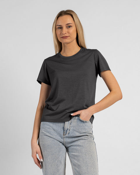 A studio portrait of a blonde woman standing and looking directly at the camera, with her left hand casually tucked into the front pocket of her light-wash blue jeans. She is wearing a regular fit, charcoal grey short-sleeved crew-neck t-shirt against a solid light grey background.