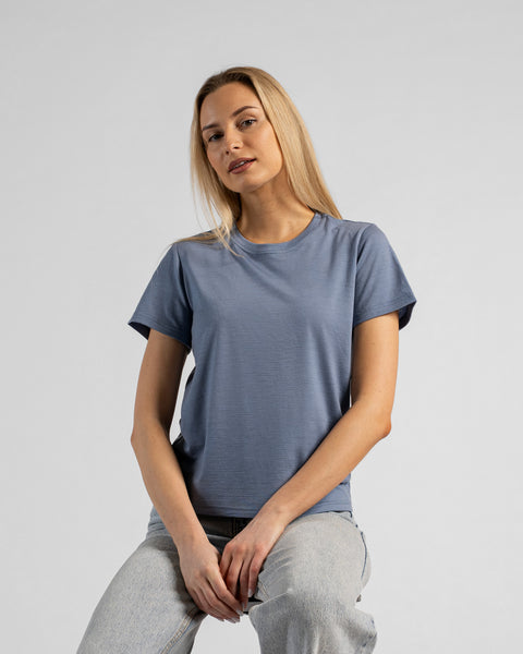A studio portrait of a blonde woman sitting and leaning slightly forward, with her hands resting on her lap. She is wearing a regular fit, dusty blue short-sleeved crew-neck t-shirt and light-wash blue jeans. The background is a solid light grey.