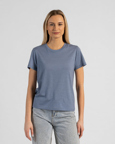 A studio portrait of a blonde woman standing and looking directly at the camera with a subtle smile. Her arms are relaxed by her sides. She is wearing a regular fit, dusty blue short-sleeved crew-neck t-shirt and light-wash blue jeans against a plain light grey background.