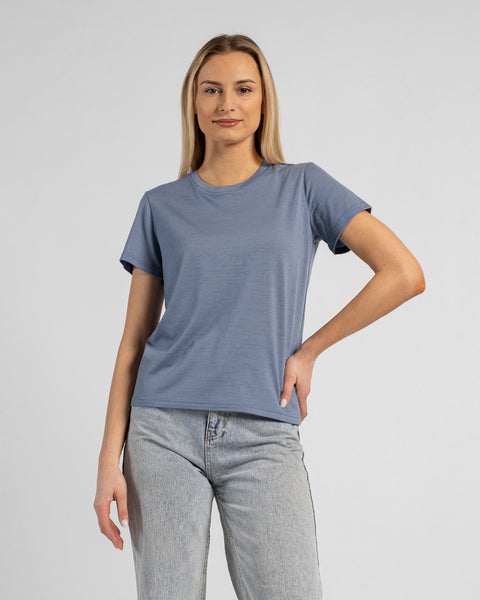A studio portrait of a blonde woman standing and looking directly at the camera with a subtle smile. She is wearing a dusty blue short-sleeved crew neck t-shirt and light wash denim jeans, with one hand resting on her hip against a plain light grey background.