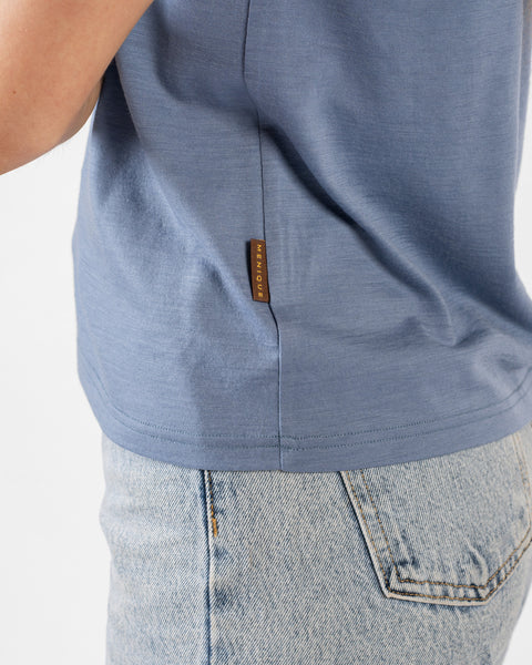 A close-up studio shot focusing on the lower side seam of a dusty blue short-sleeved t-shirt worn by a woman, highlighting a small brown "MENIQUE" logo tag. She is also wearing light wash denim jeans.
