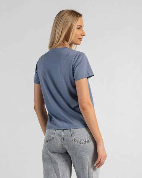 A back view studio portrait of a blonde woman standing against a plain light grey background. She is wearing a dusty blue short-sleeved crew neck t-shirt and light wash denim jeans, with her head turned to look off to her right.