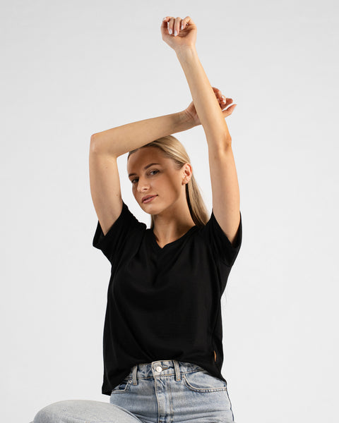 A studio portrait of a blonde woman looking directly at the camera. She is wearing a black short-sleeved V-neck t-shirt and light wash denim jeans, with her arms gracefully raised and hands resting together above her head against a plain light grey background.