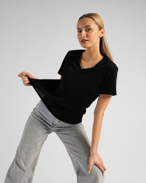 A studio portrait of a blonde woman leaning slightly forward, playfully pulling the fabric of her black t-shirt with her right hand. Her left hand is resting on her knee. She is wearing a regular fit, solid black short-sleeved v-neck t-shirt and light-wash blue jeans against a plain light grey background.