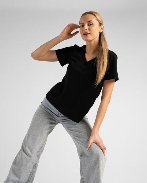 A studio portrait of a blonde woman leaning forward and looking directly at the camera. She is wearing a black short-sleeved V-neck t-shirt and light wash denim jeans, with one hand raised near her temple and the other resting on her knee against a plain light grey background.