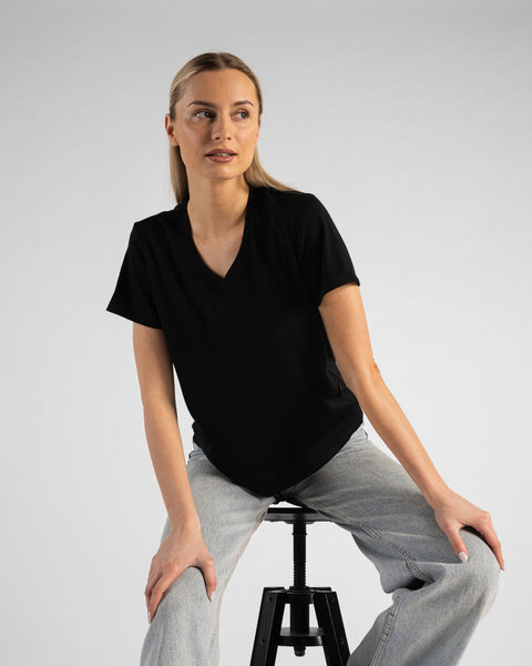 A studio portrait of a blonde woman sitting on a black stool and looking off to her right. She is wearing a black short-sleeved V-neck t-shirt and light wash denim jeans, with both hands resting lightly on her knees against a plain light grey background.