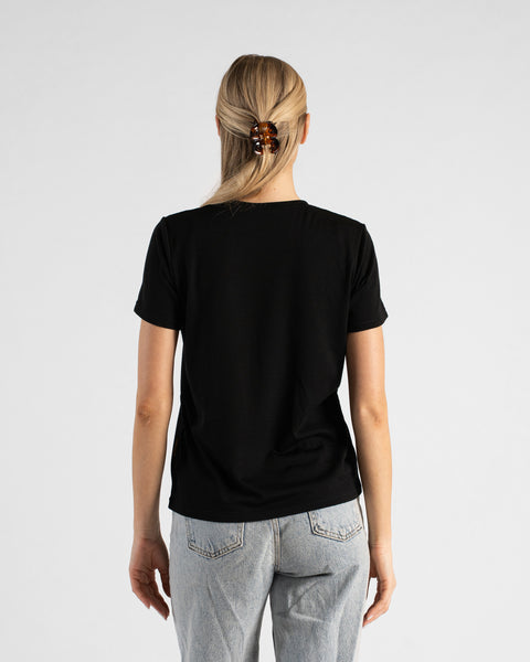A back view studio shot of a blonde woman standing against a plain light grey background. She is wearing a black short-sleeved V-neck t-shirt and light wash denim jeans, with her hair styled half-up using a brown tortoiseshell claw clip.