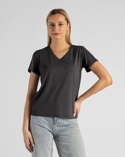 A studio portrait of a blonde woman standing and looking directly at the camera, with her left hand resting on her hip. She is wearing a regular fit, charcoal grey short-sleeved v-neck t-shirt and light-wash blue jeans against a plain light grey background.