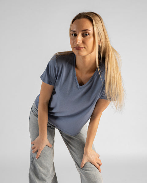 A studio portrait of a blonde woman leaning forward and looking directly at the camera. Her hands are resting on her thighs just above her knees. She is wearing a regular fit, dusty blue short-sleeved v-neck t-shirt and light-wash blue jeans against a plain light grey background.