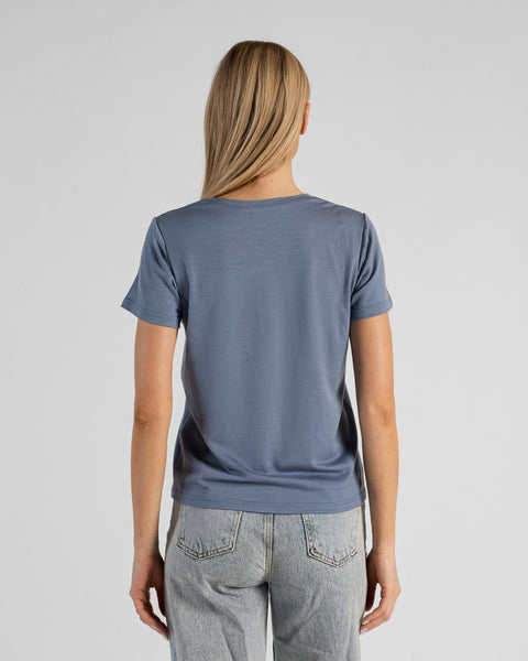 A back view studio shot of a blonde woman standing against a plain light grey background. She is wearing a dusty blue short-sleeved t-shirt and light wash denim jeans, with her straight blonde hair falling down her back.