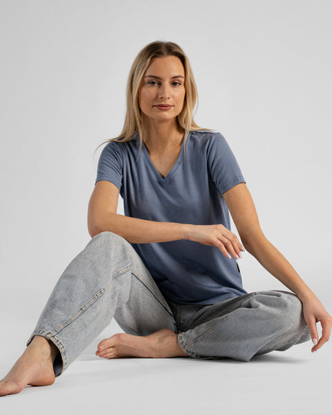 A studio portrait of a barefoot blonde woman sitting comfortably on the floor with her legs loosely crossed and bent. She is looking directly at the camera, wearing a dusty blue short-sleeved V-neck t-shirt and light wash denim jeans against a plain light grey background.