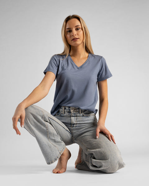 A studio shot of a barefoot blonde woman in a low crouch or kneeling pose on the floor, looking directly at the camera. She is wearing a dusty blue short-sleeved V-neck t-shirt and wide-leg light wash denim jeans, with her hands resting lightly on her knees against a plain light grey background.