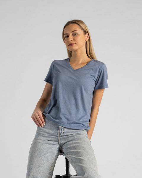 A studio portrait of a blonde woman sitting on a stool and looking directly at the camera. She is leaning back slightly, with her hands resting on her lap. She is wearing a regular fit, dusty blue short-sleeved v-neck t-shirt and light-wash blue jeans against a plain light grey background.