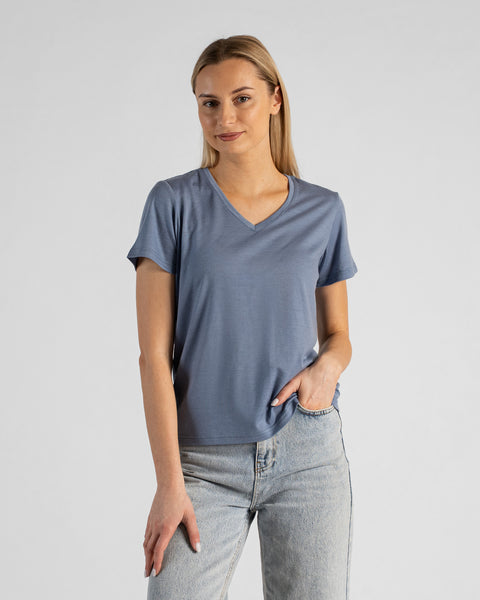 A studio portrait of a blonde woman standing and looking directly at the camera with a subtle smile. She is wearing a dusty blue short-sleeved V-neck t-shirt and light wash denim jeans, with one hand casually resting in her front pocket against a plain light grey background.