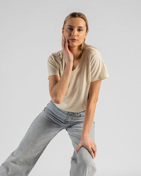 A studio portrait of a blonde woman leaning forward and looking directly at the camera. Her right hand is gently touching her cheek, and her left hand is resting on her knee. She is wearing a regular fit, light cream natural-colored short-sleeved v-neck t-shirt and light-wash blue jeans. The background is a solid light grey.