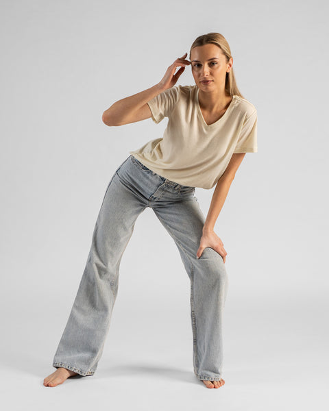 A studio shot of a barefoot blonde woman standing with a wide stance and leaning forward, resting one hand on her knee and bringing the other up to her temple. She is wearing a natural off-white short-sleeved V-neck t-shirt and wide-leg light wash denim jeans against a plain light grey background.