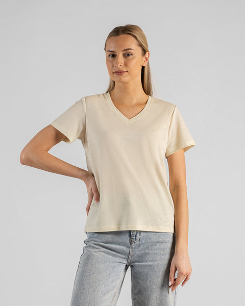 A studio portrait of a blonde woman standing with one hand resting on her hip, looking directly at the camera with a subtle smile. She is wearing a natural off-white short-sleeved V-neck t-shirt and light wash denim jeans against a plain light grey background.