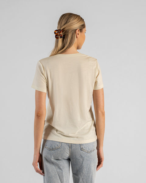 A back view studio shot of a blonde woman standing against a plain light grey background. She is wearing a natural off-white short-sleeved V-neck t-shirt and light wash denim jeans, with her hair styled half-up using a brown tortoiseshell claw clip.