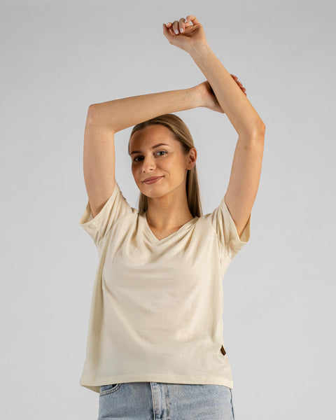 A studio portrait of a blonde woman standing with her arms gracefully crossed above her head, smiling at the camera. She is wearing a natural off-white short-sleeved V-neck t-shirt and light wash denim jeans against a plain light grey background.
