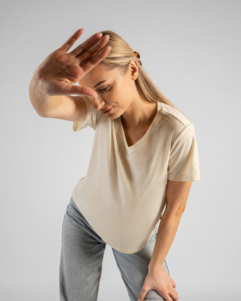 A studio portrait of a blonde woman playfully leaning forward and holding one hand up to shield her face from the camera. She is wearing a natural off-white short-sleeved V-neck t-shirt and light wash denim jeans against a plain light grey background.